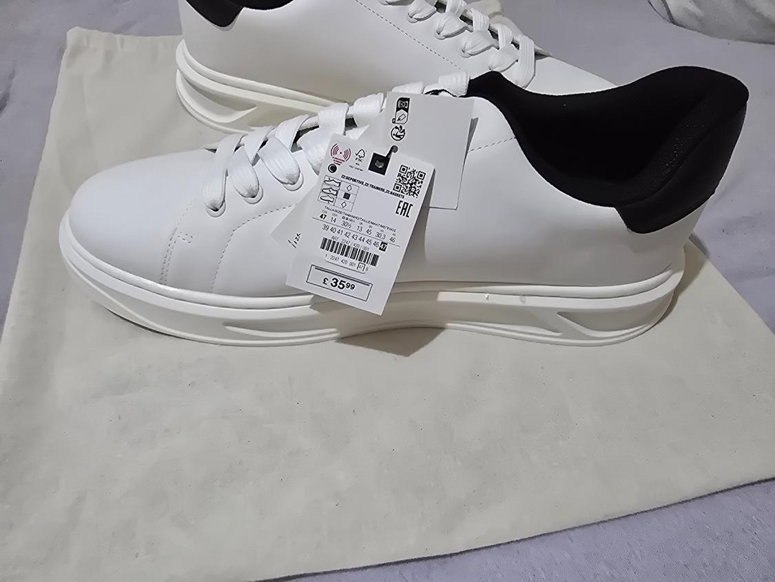 White trainers with black heels on a cloth bag. They have a label bearing the text EAC and £35.99.