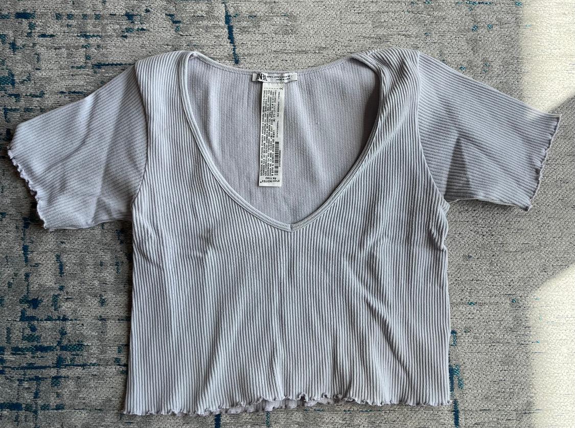 Light grey ribbed T-shirt with a V-neck and short sleeves, on a grey rug with blue accents. Visible label on the collar.