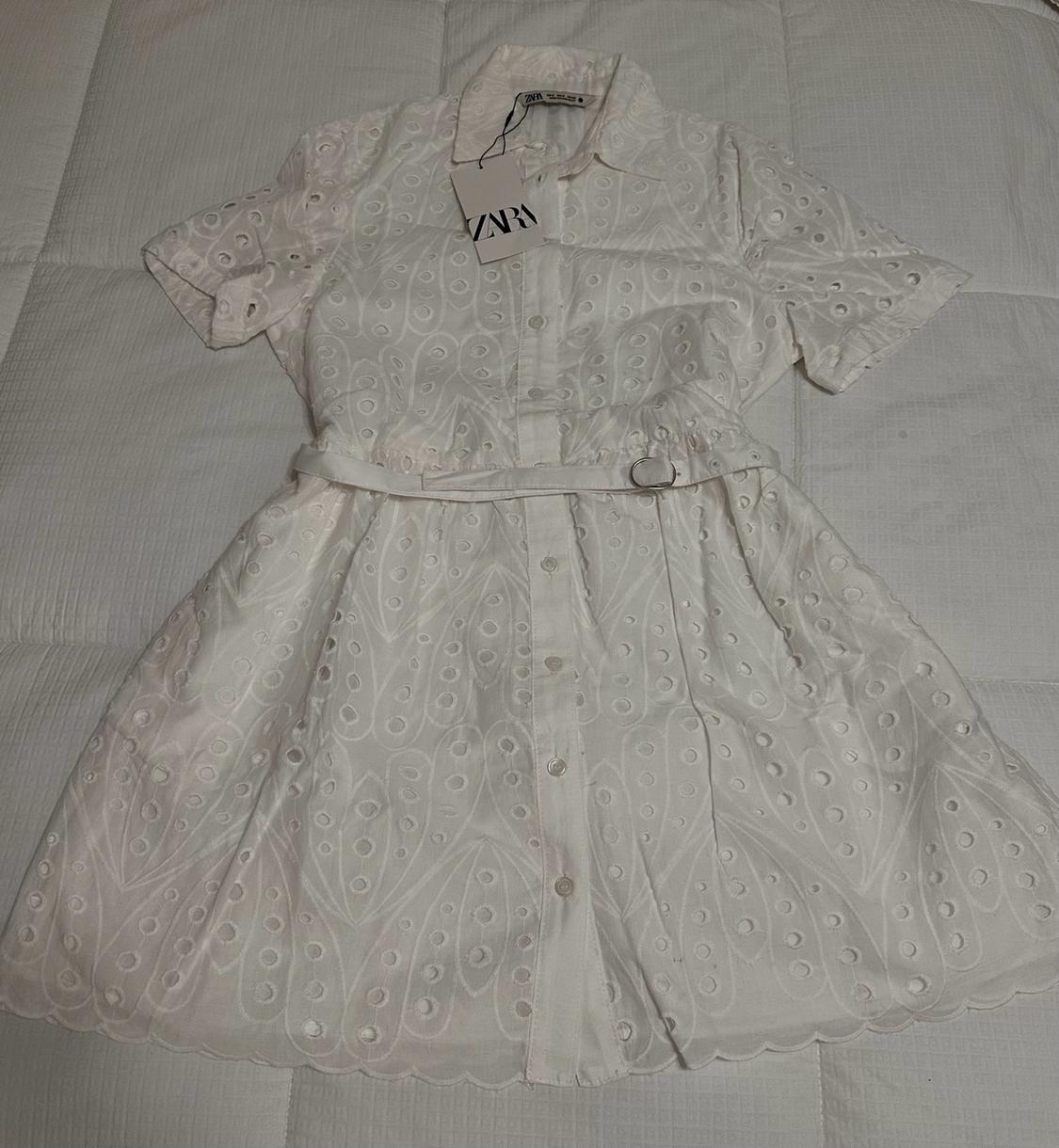 White short-sleeved dress with eyelet embroidery, buttons, and a belt, against a quilted background. ZARA label.