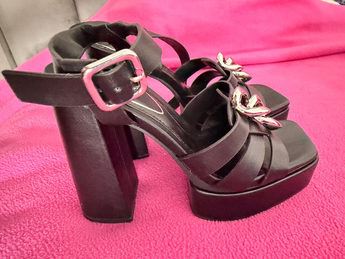 Black high-heeled sandals with silver buckle and metallic embellishment on the upper, on a pink fabric background.