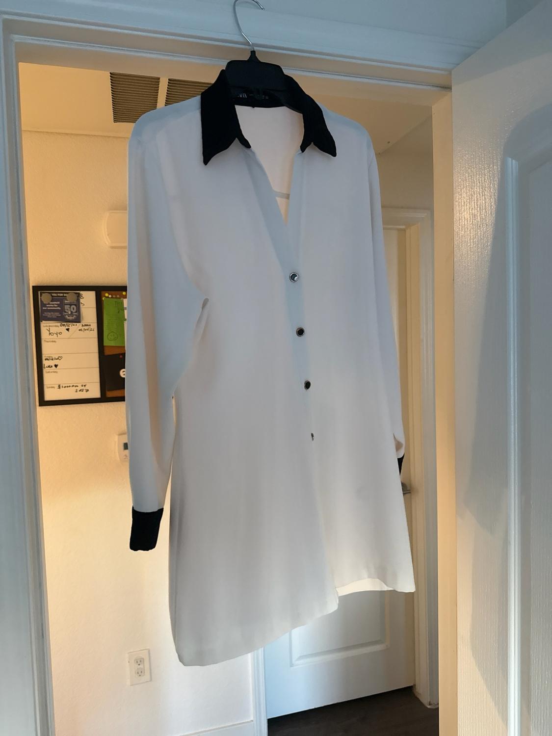 White long-sleeved shirt with a collar and black cuffs, hanging on a coat hanger in the frame of an open door in a domestic setting.