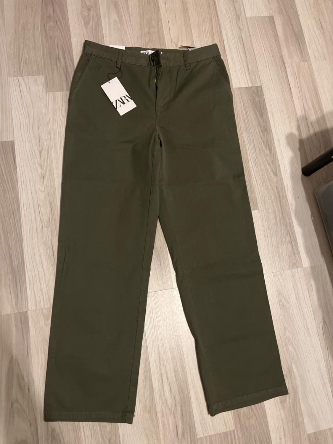 Green straight-cut trousers on a light wooden floor, with a hanging tag that says "ZARA".