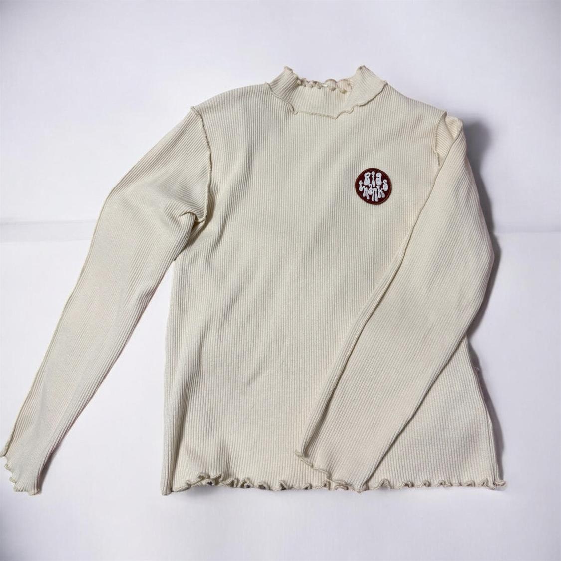 Beige knitted jumper with a high neck and ruffled edges. Features a red circular patch with white text on the chest.