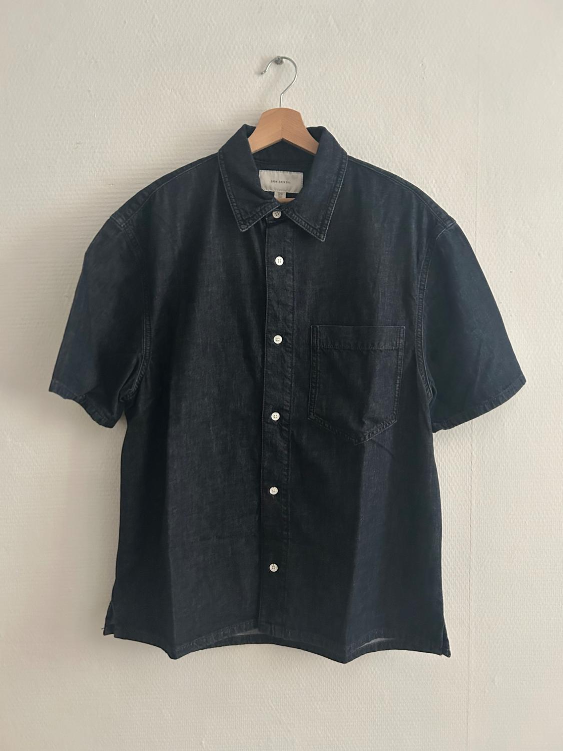 Black short-sleeved denim shirt hanging on a wooden hanger against a white wall. White buttons visible.