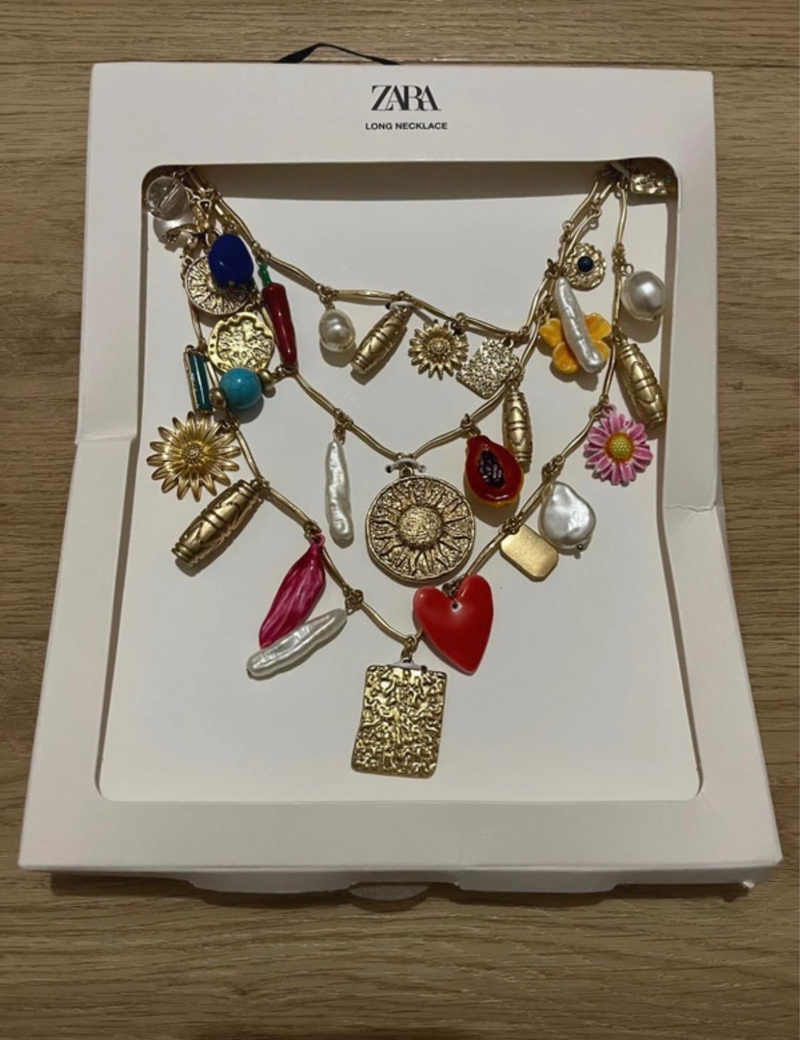 Long Zara necklace with gold pendants, pearls, flowers, and a red heart, presented in a white box on a wooden surface.