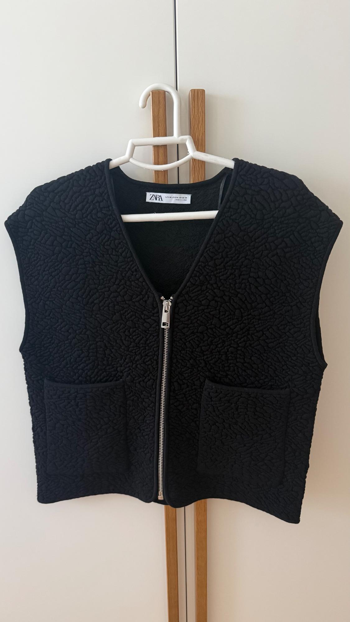 Textured black gilet with a front zip and pockets, hanging on a hanger against a white background. Label with the text ZARA.