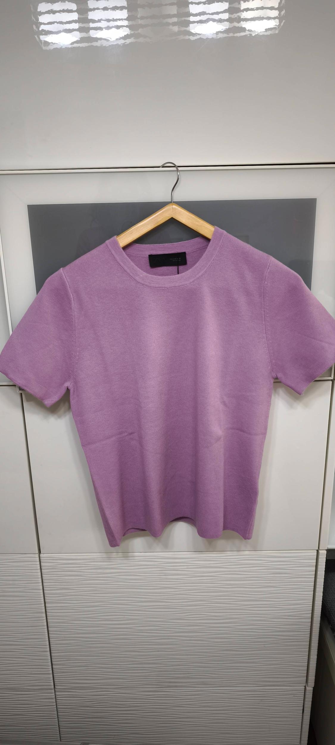 Lilac short-sleeved T-shirt hanging on a wooden hanger against a white and grey background.