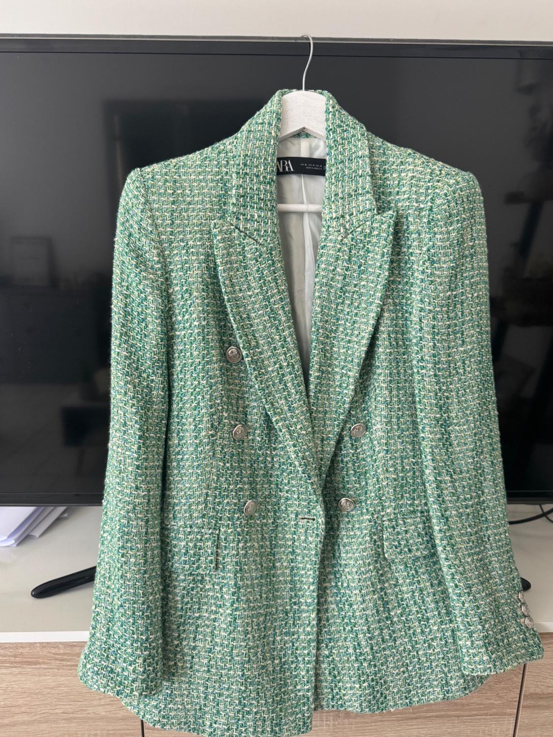 Green textured fabric jacket with silver buttons, hanging on a hanger against a black background. Label visible.