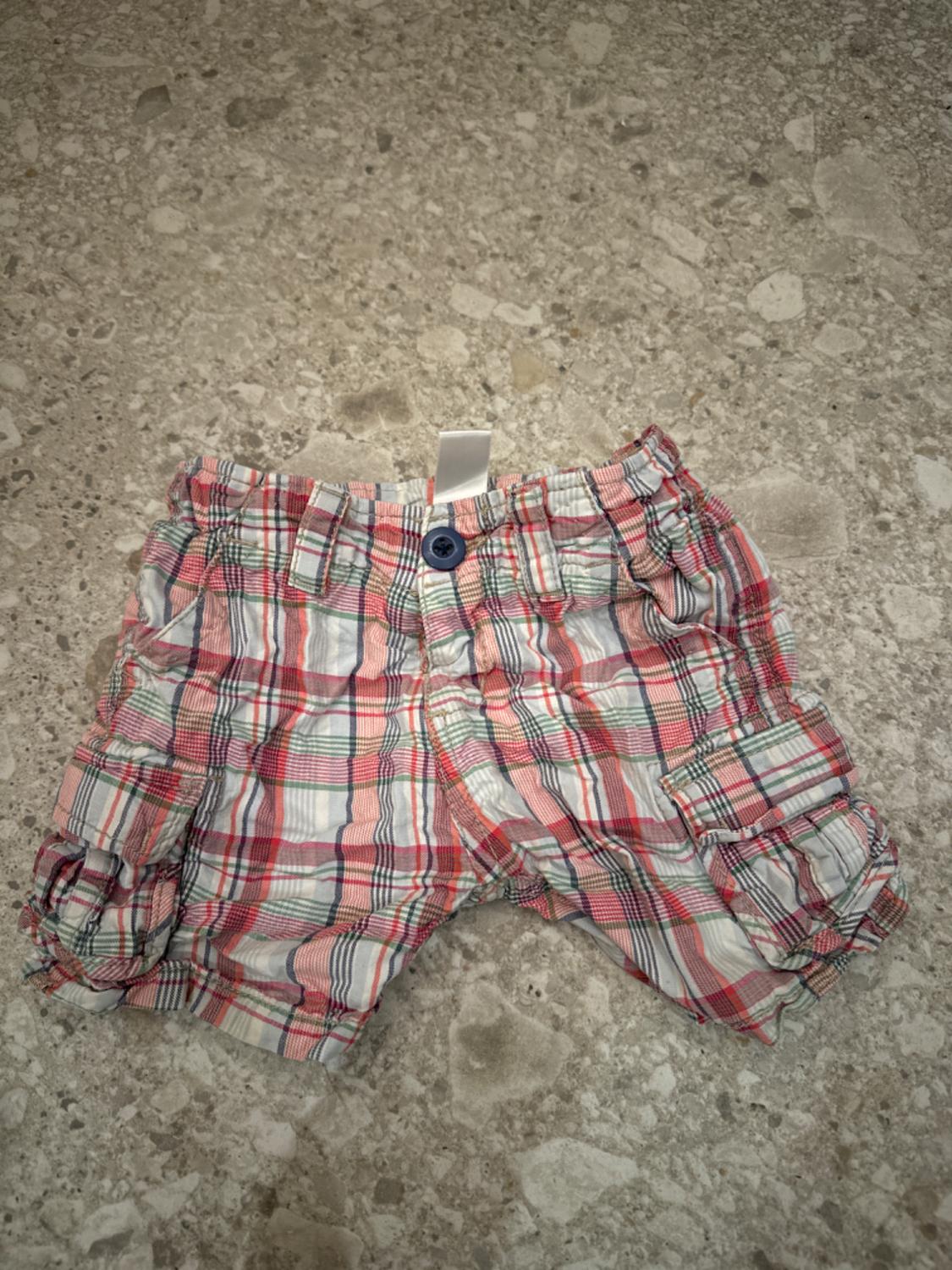 Red, white, and green checked shorts with a blue button, on a grey stone floor.