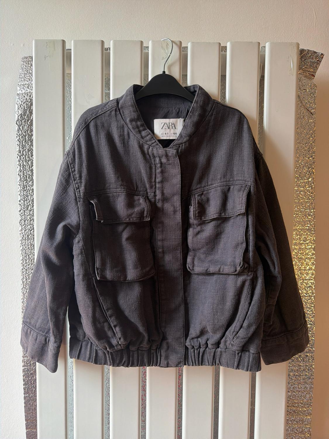 Dark grey jacket with pockets and an elasticated hem, hanging on a white radiator against a shiny silver background. Text: ZARA.