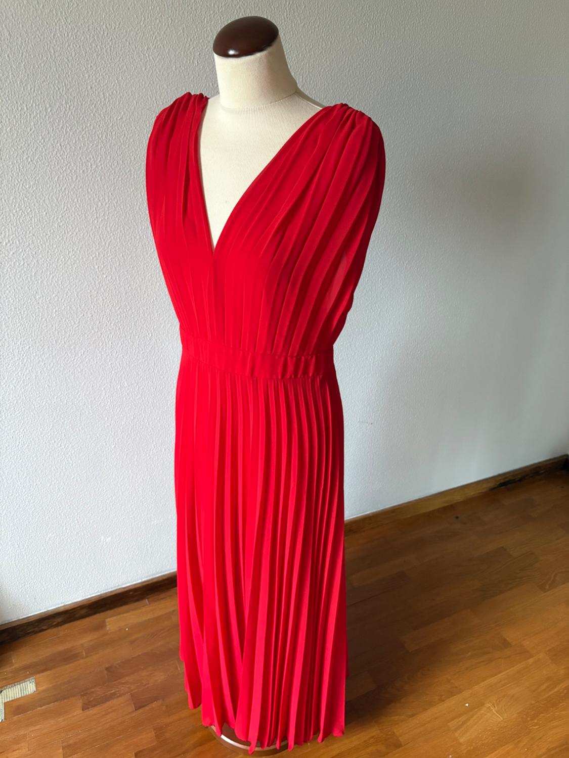 Red, sleeveless, pleated maxi dress with a deep V-neckline, displayed on a mannequin on a wooden floor in front of a white wall.
