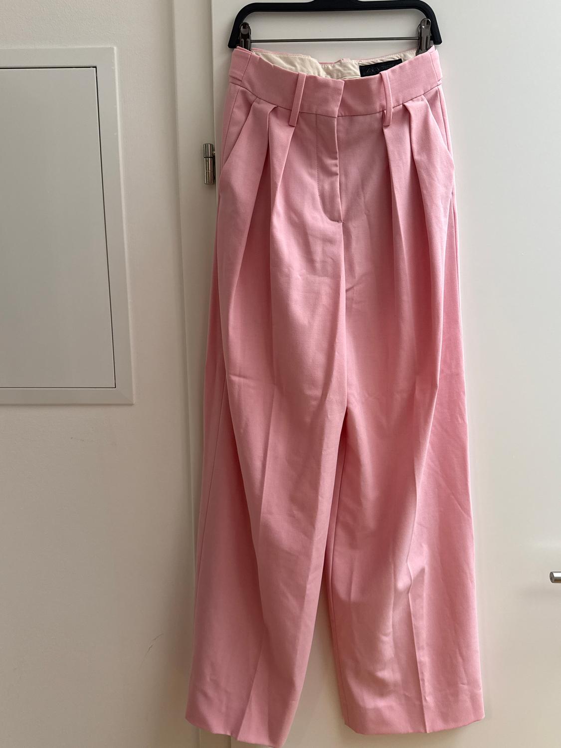 Light pink trousers hanging on a black hanger over a white door. Pleated design with a high waist.