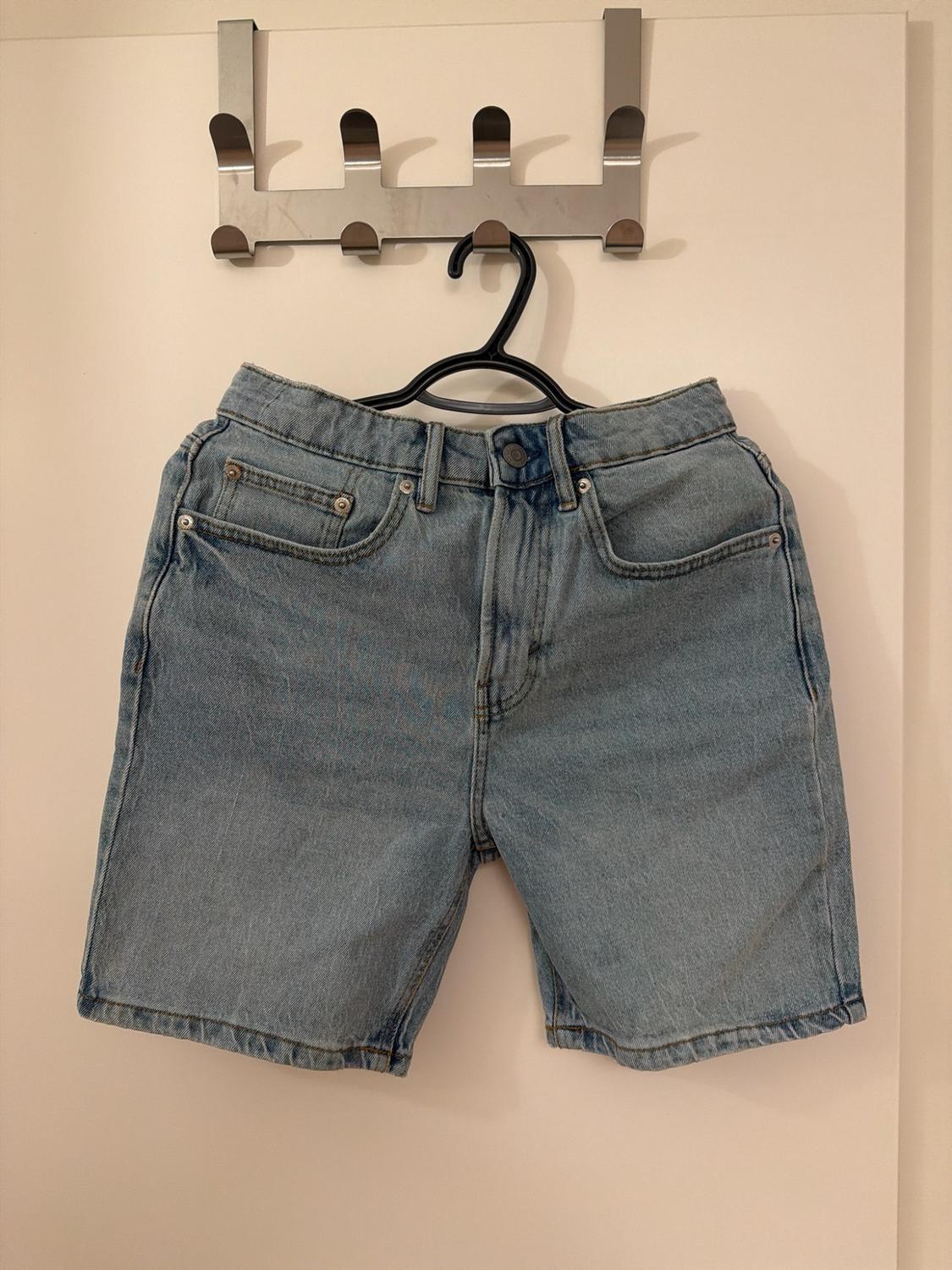 Light blue denim shorts hanging on a black hanger on a metal coat rack on a white door.