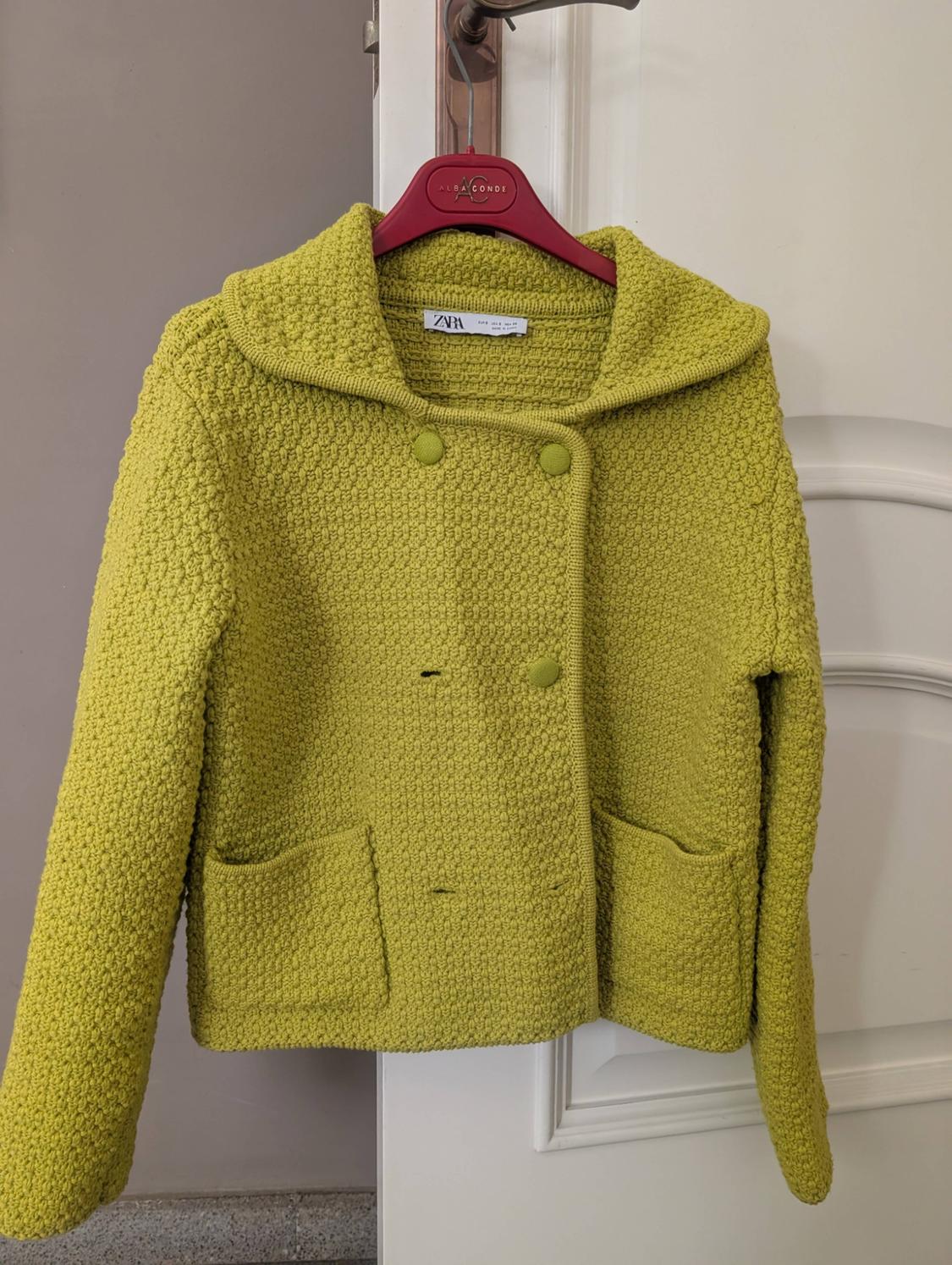 Green knitted cardigan with large buttons and front pockets, hanging on a red hanger with the label "Zara".