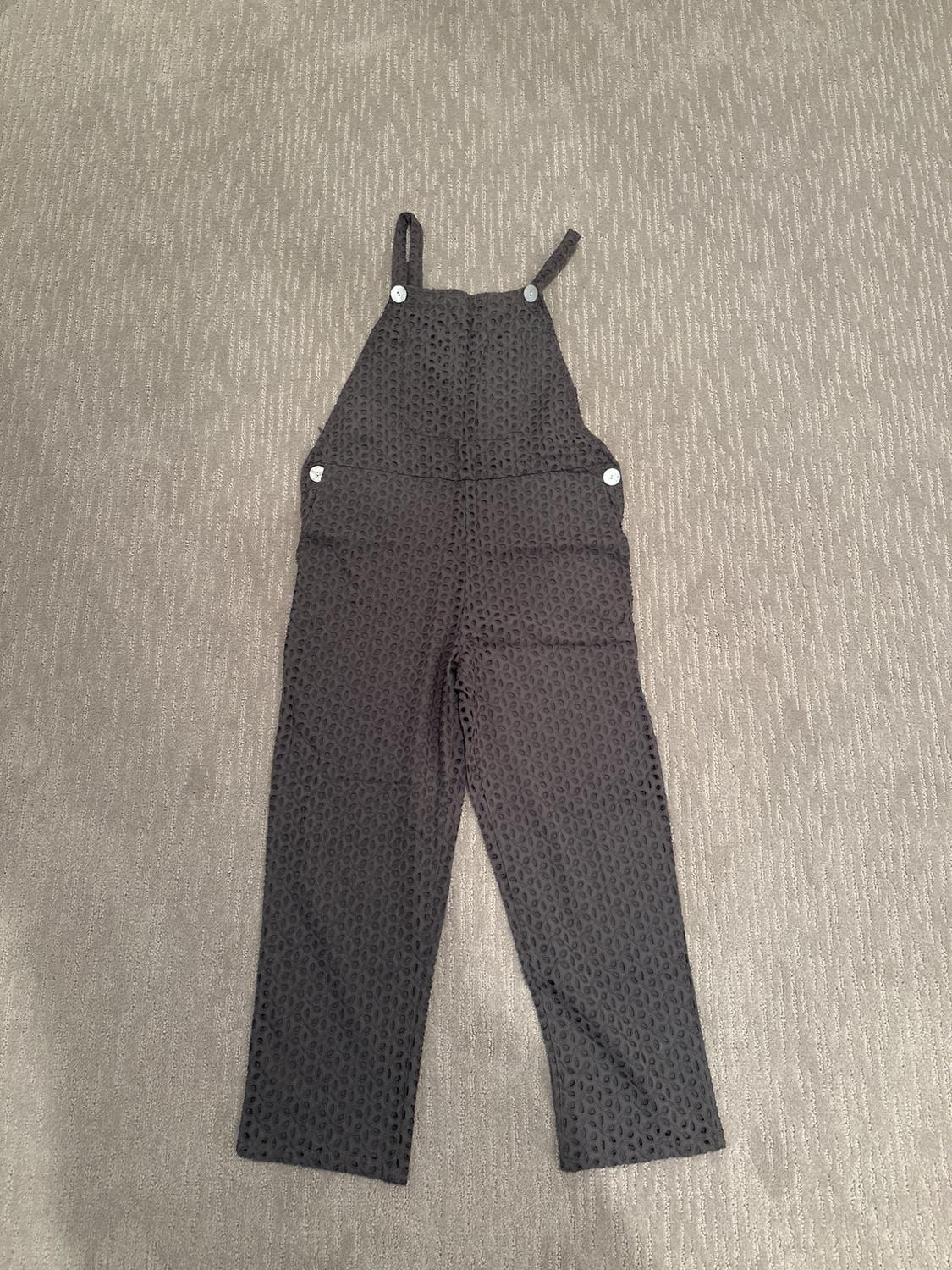 Dark gray children's jumpsuit with white straps and buttons, on a textured beige fabric background.