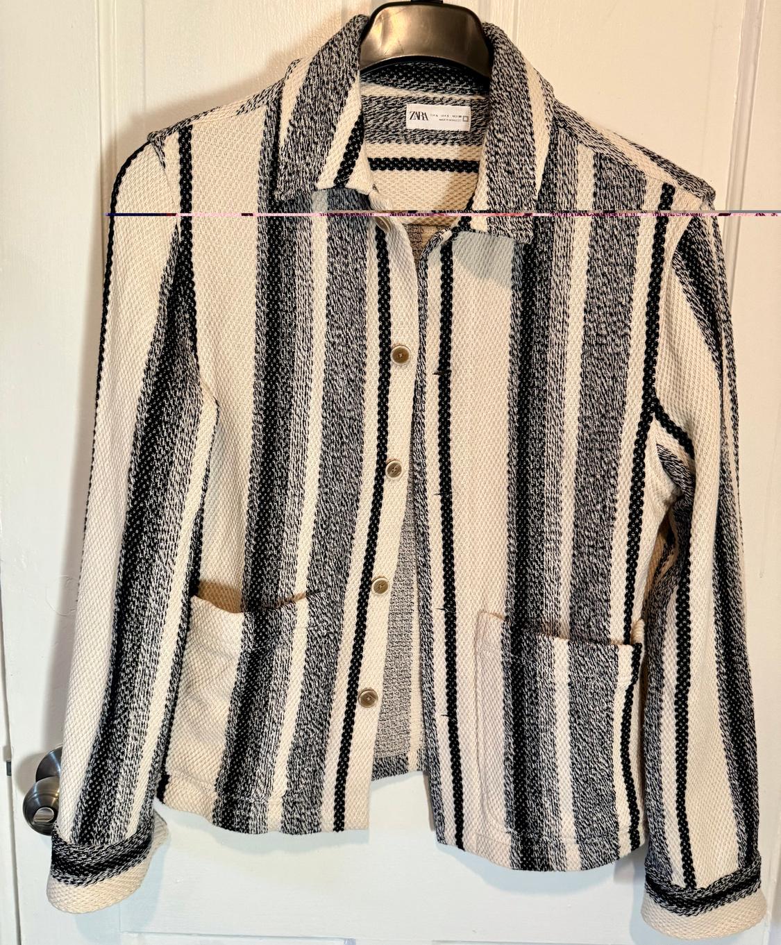 Black and white vertical striped jacket hanging on a hanger. It has front buttons and two large pockets.