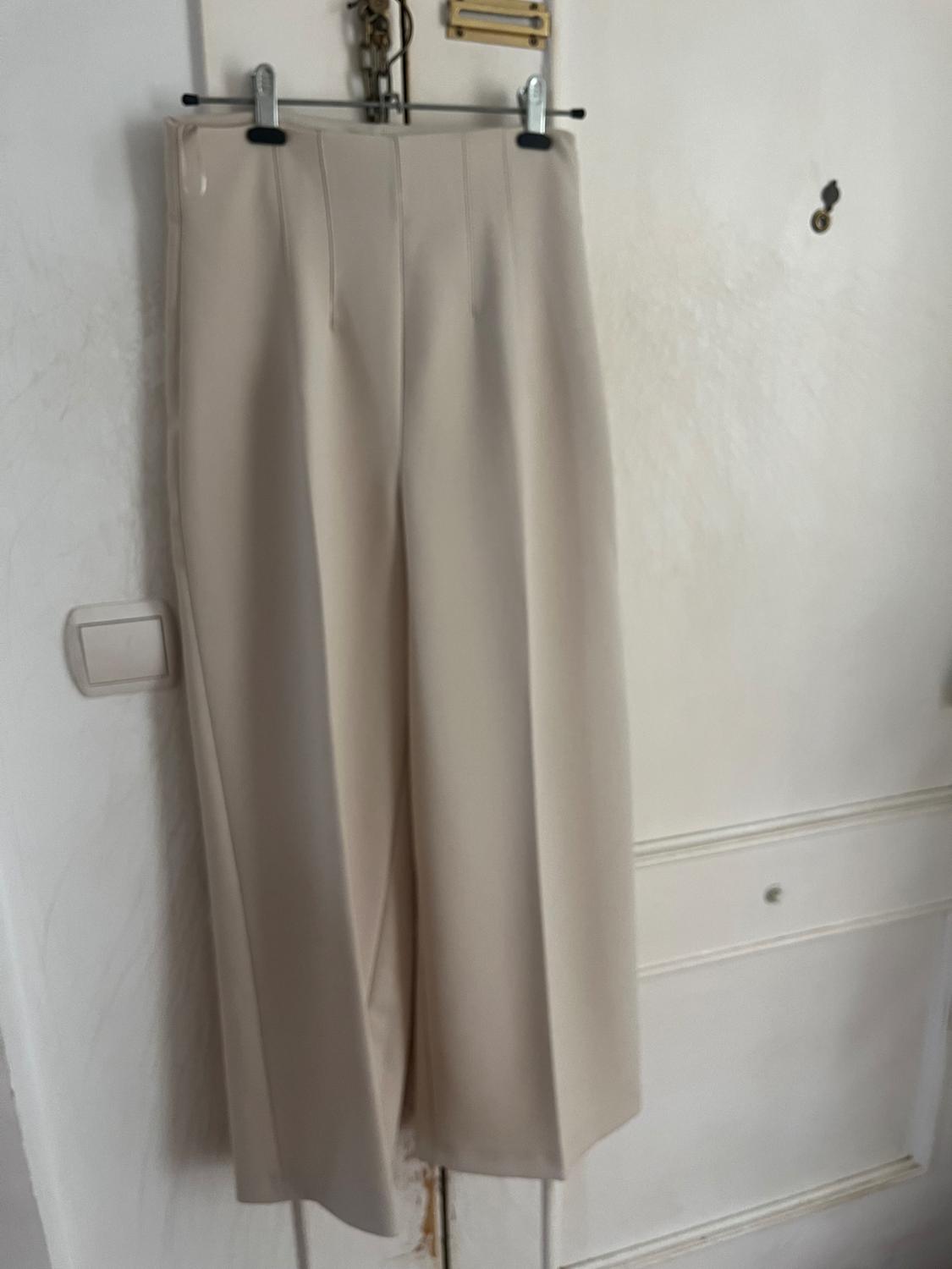 Beige high-waisted wide-leg trousers with front seams, hanging on a hanger on a white interior door. Casual and relaxed style.