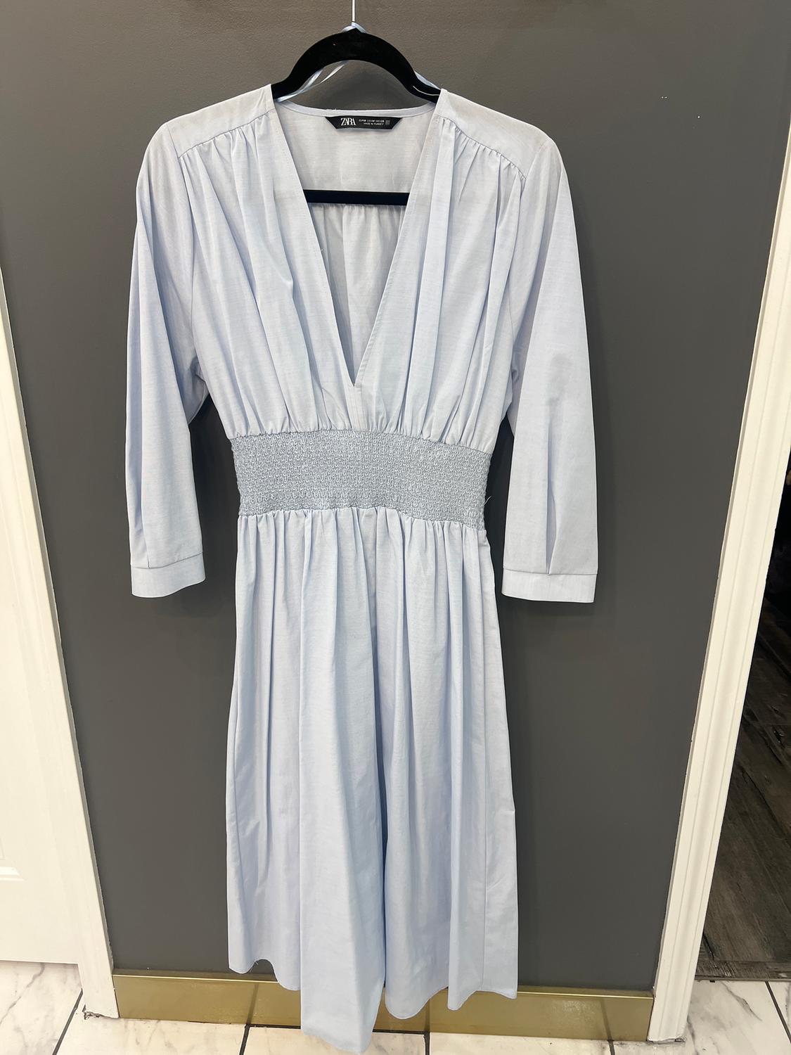 Light blue V-neck dress with ruched waist, hanging on a black hanger against a gray wall.