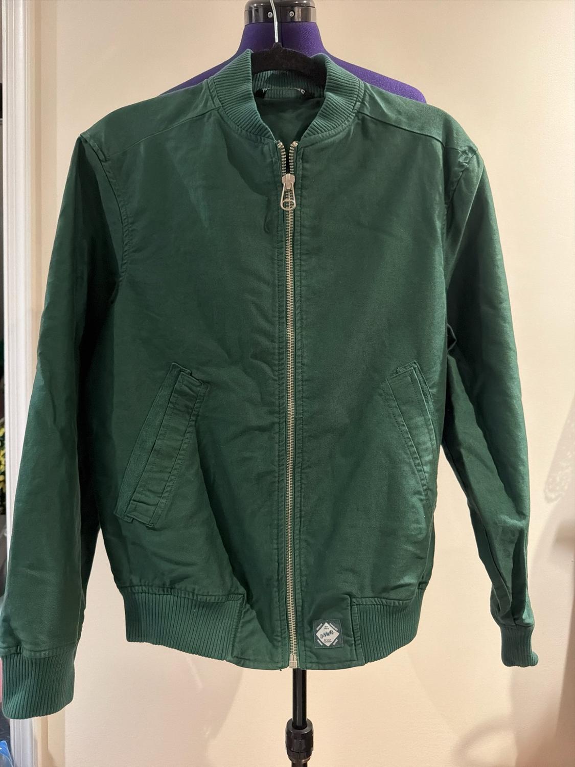 Green bomber jacket on a mannequin. Design with a zip fastening, front pockets and a patch at the hem bearing the text “DNWR”.