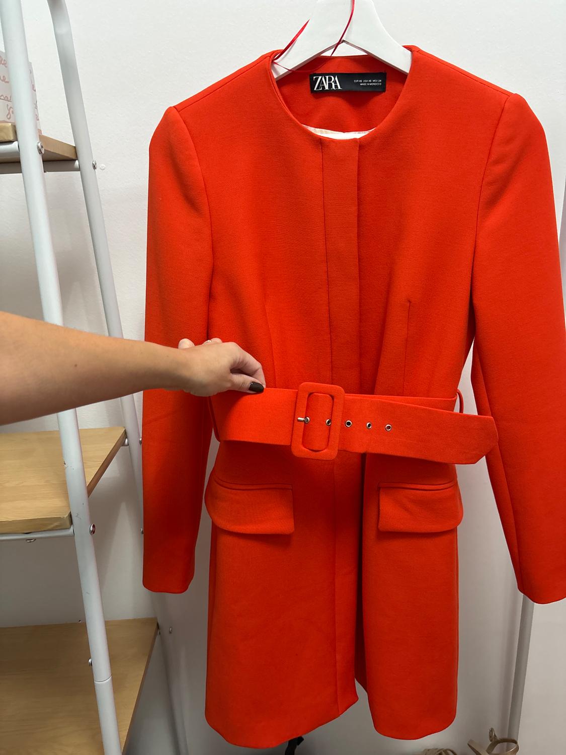 A red coat with a belt and pockets is hanging on a white hanger. A hand is holding the belt. The visible label reads: ZARA.