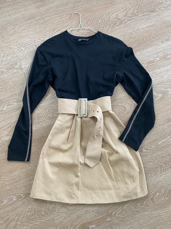 Black long-sleeved dress with a wide beige belt on a wooden floor. Stitching detail on the sleeves.