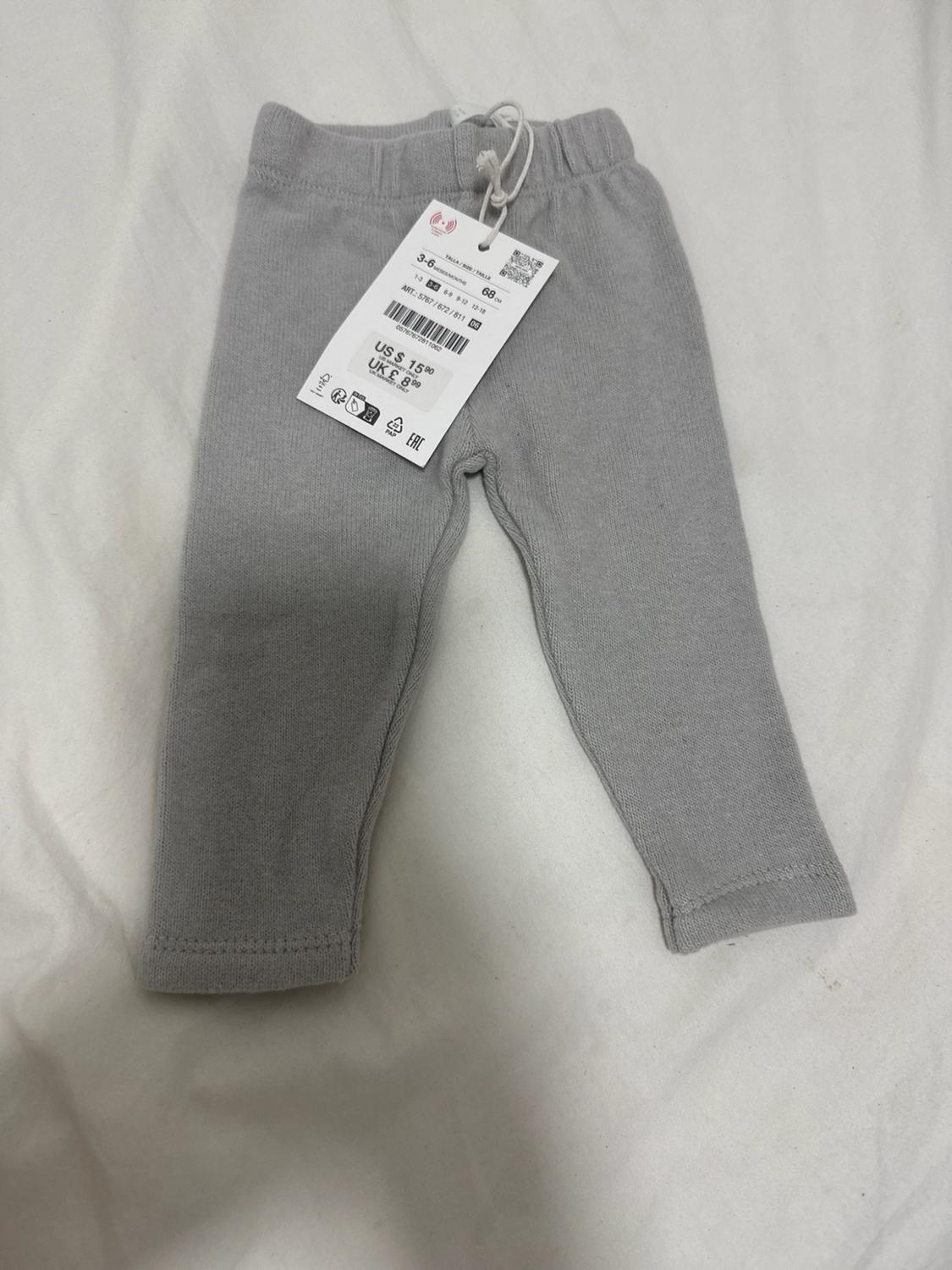 Gray knit baby pants on white fabric. Tag: 3-6, 68 cm, ART.: 5767/672/811, US $ 15.90, UK £ 8.99.