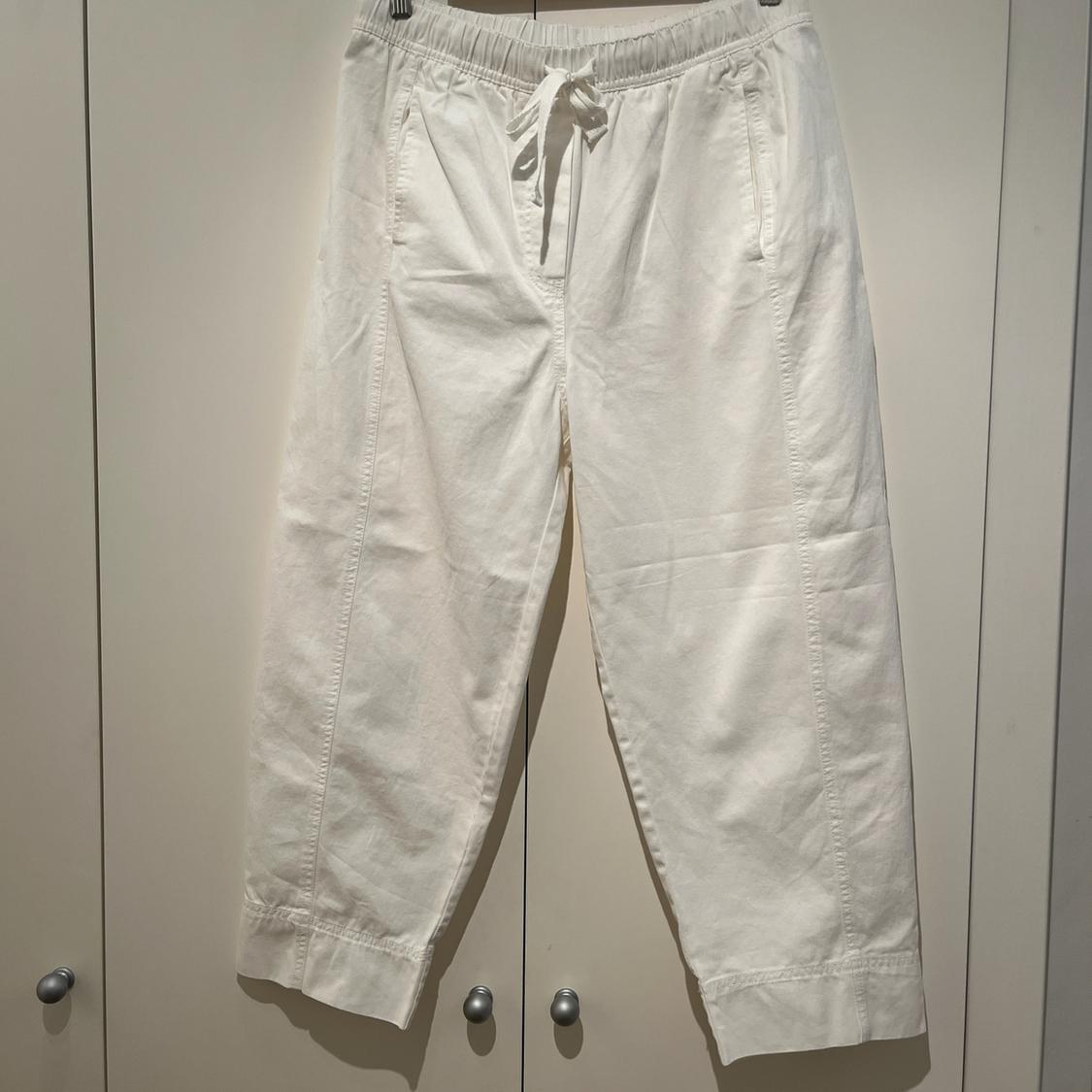 Loose-fit white trousers with an elasticated waist, drawstring and front seams, hanging in front of a light-coloured wardrobe with silver knobs.