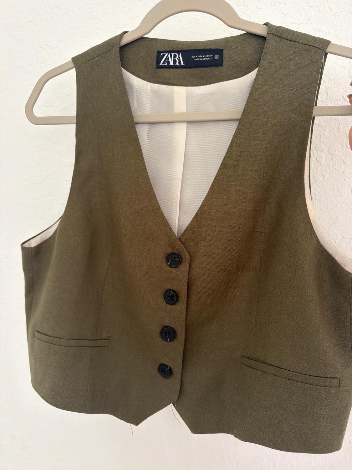 Olive green button-up waistcoat hanging on a coat hanger against a white background. Text: ZARA EUR M USA M MEX 28 MADE IN MOROCCO.