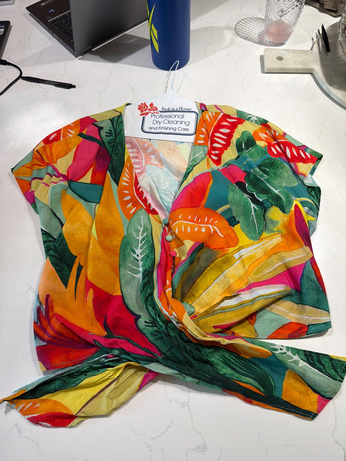 Multicolor wrap blouse with tropical print on marble. Text: Fresh as a Flower Professional Dry Cleaning and Finishing Care.