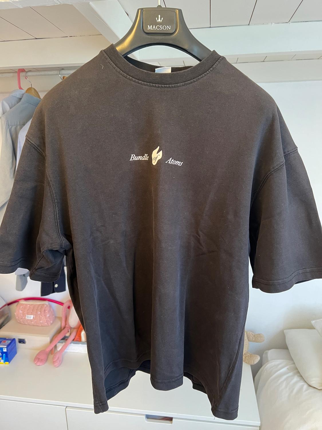 Black T-shirt hanging on a Macson hanger, with "Bundle Atoms" text and a flame design on the chest. Bedroom background.