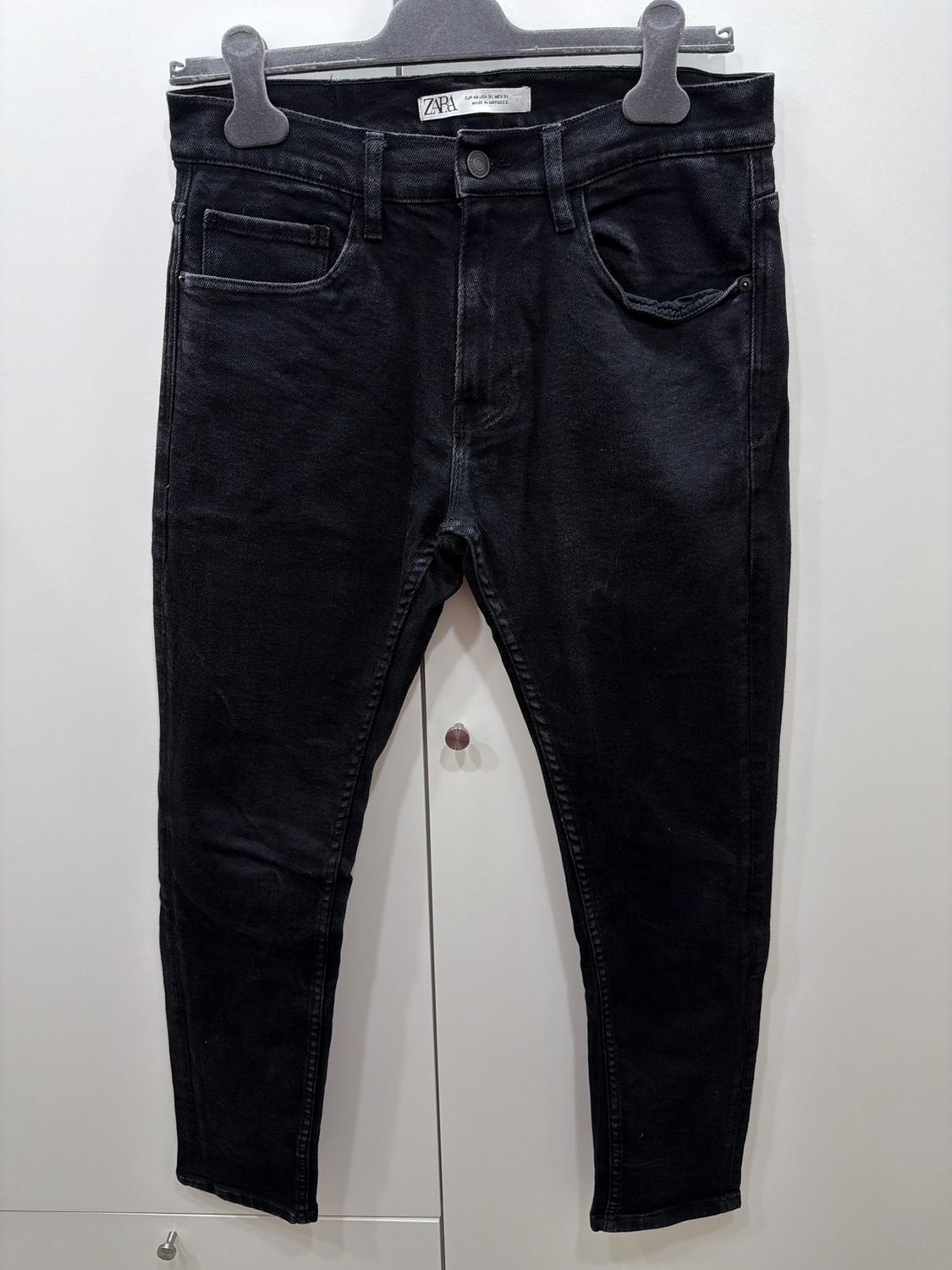 Black denim trousers hanging on a grey hanger, with a visible label that reads "Zara". White wardrobe background.