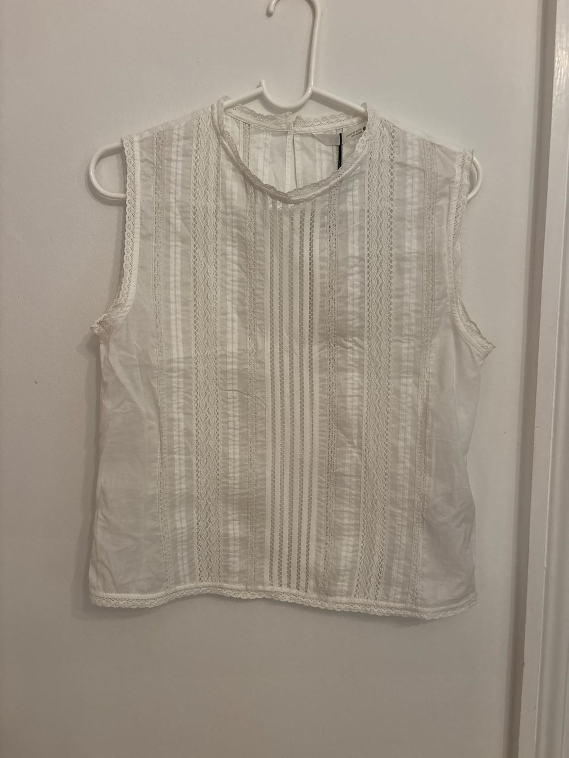 White sleeveless blouse with vertical lace details and a high neck, hanging on a white hanger against a white wall background.