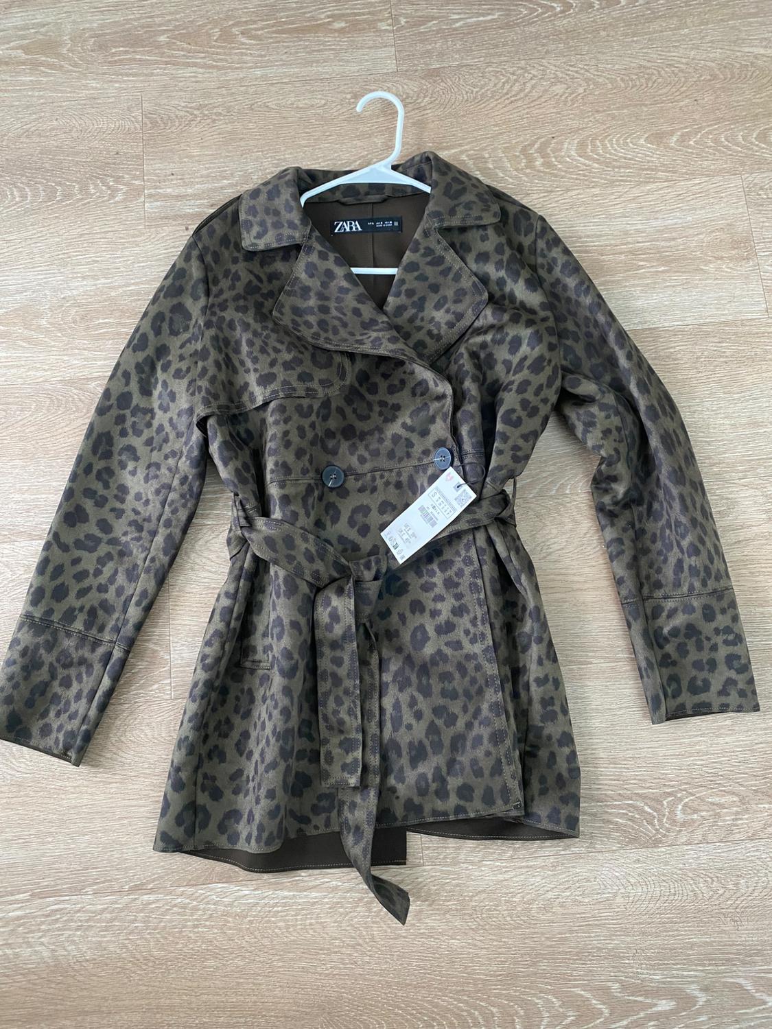 Brown leopard print coat with belt and buttons, hanging on a white hanger over light wood flooring. Visible tag.