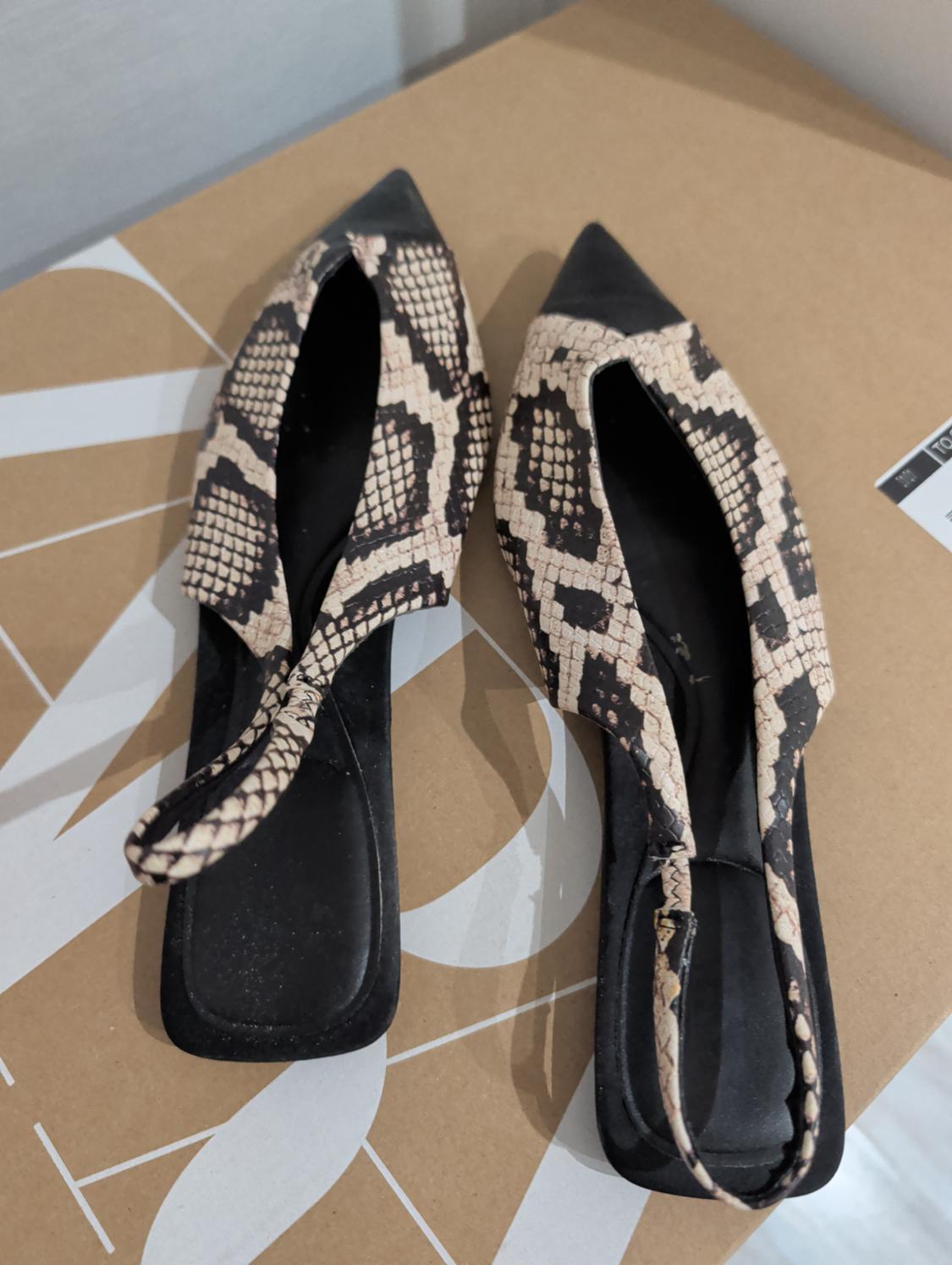 Flat mules με animal print φιδιού και μαύρη μύτη, πάνω σε ένα χάρτινο κουτί με μεγάλα λευκά γράμματα.