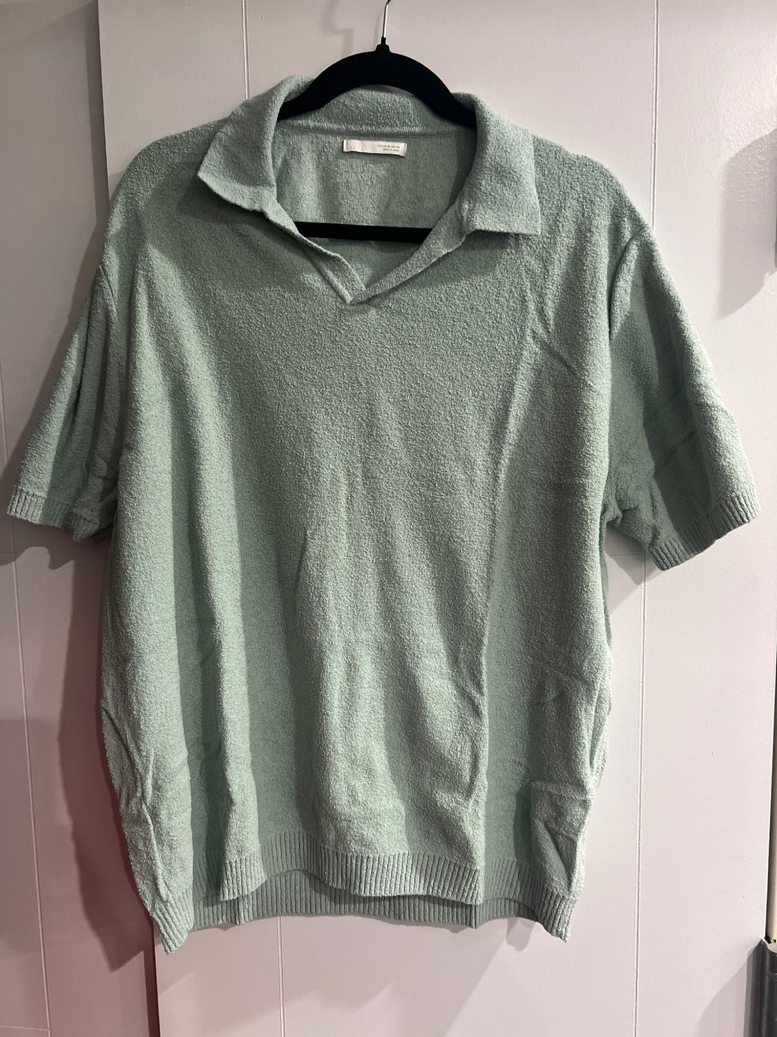 Light green short-sleeved polo shirt in a textured fabric with a V-neck, hanging on a black hanger over a white door.
