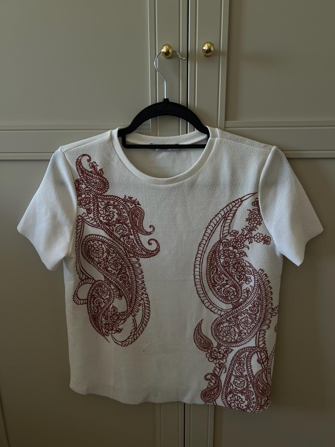 White short-sleeved T-shirt with maroon print on the sides, hanging on a black hanger in front of a light-coloured wardrobe.