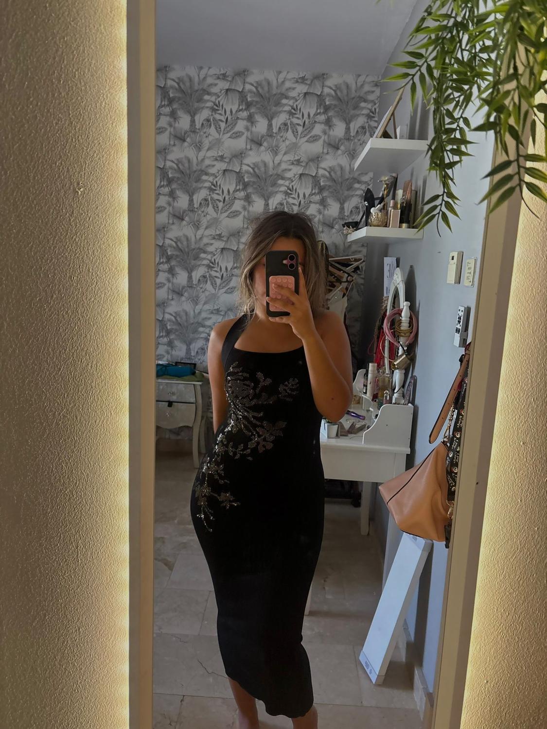 Woman taking a mirror selfie in her bedroom. She is wearing a fitted black dress with a halter neckline and sparkling floral details.