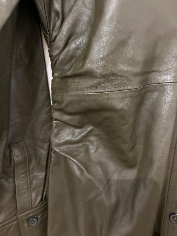 Close-up of an olive green leather jacket with a wrinkled texture and visible stitching. A dark button is visible at the bottom.
