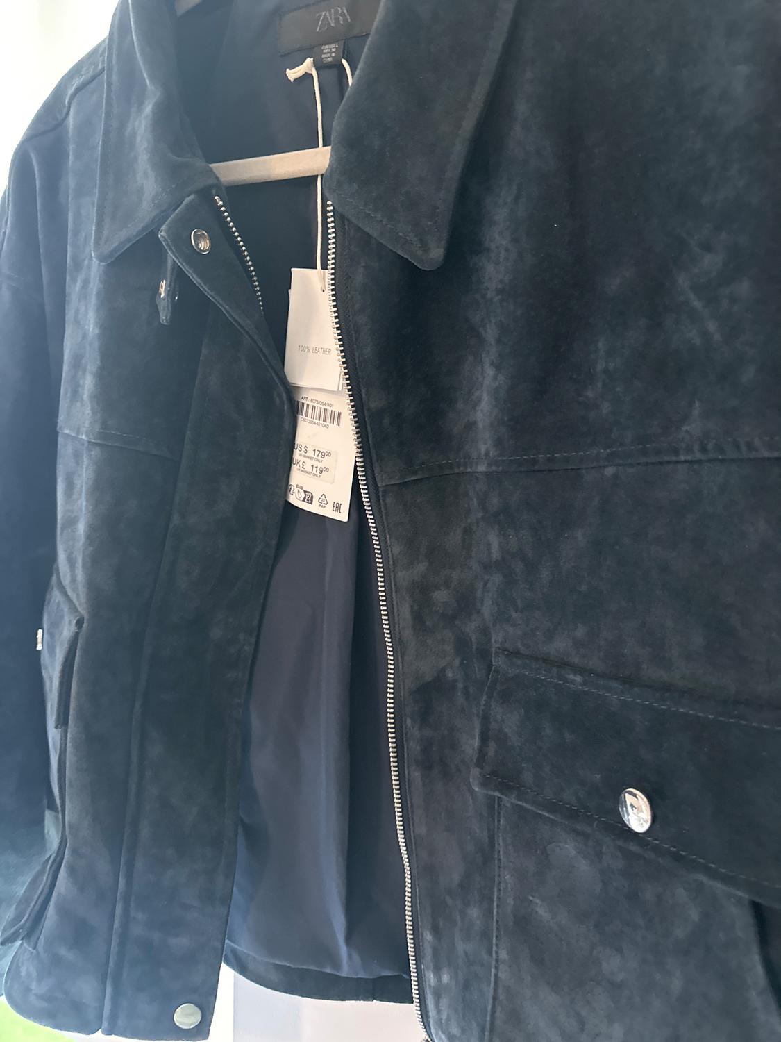 Black leather jacket hanging on a hanger, with a metal zipper and a visible tag with the text "100% LEATHER".