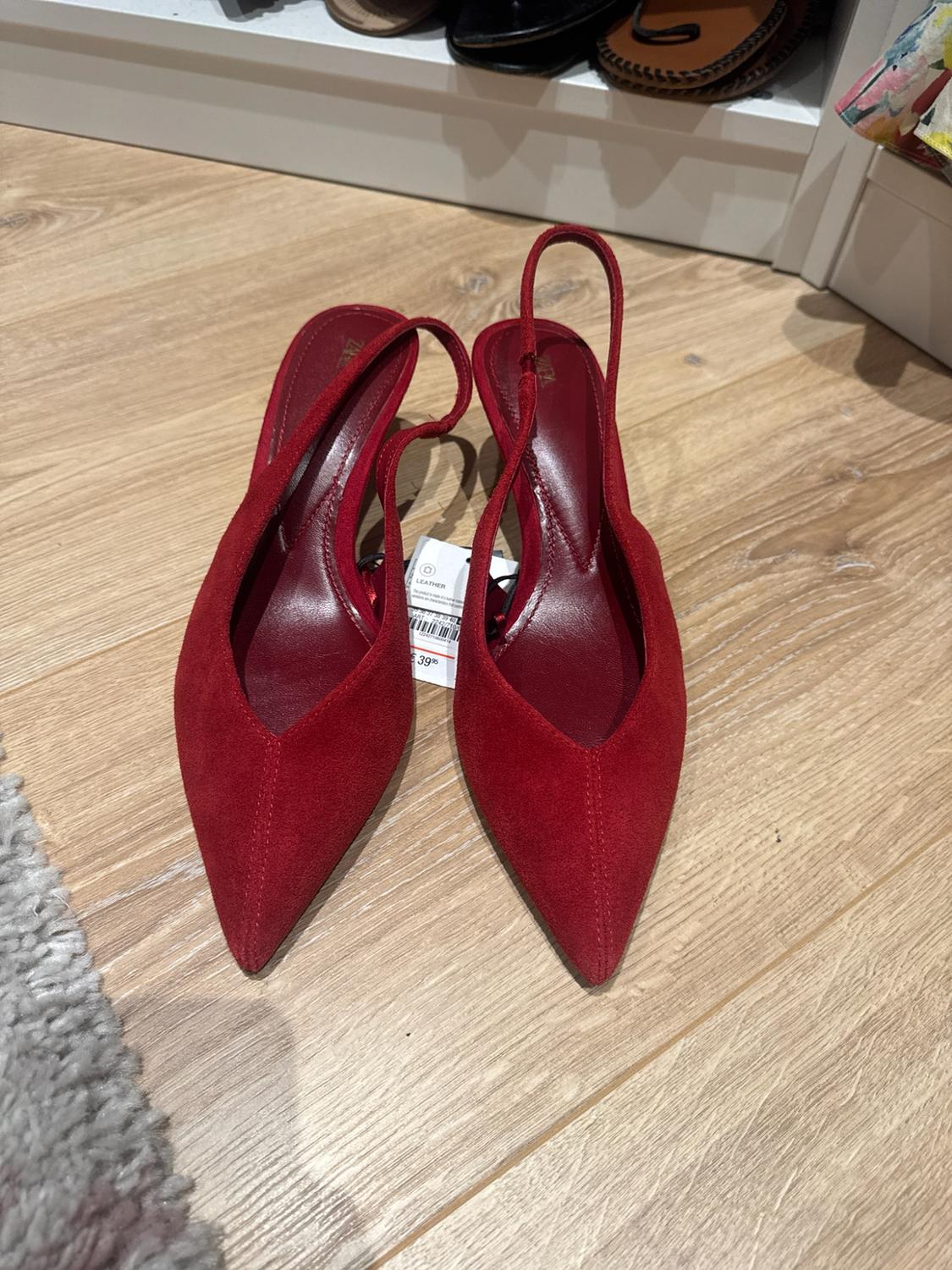 Red suede slingback shoes on a wooden floor. Label with the text LEATHER 39.95. Casual style, with a rug and shoe rack in the background.