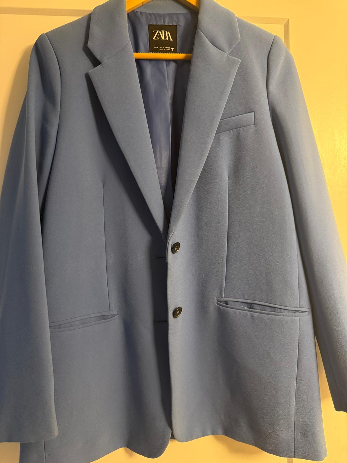 Light blue jacket hanging on a yellow hanger. Visible label with the text "ZARA". Beige door background.