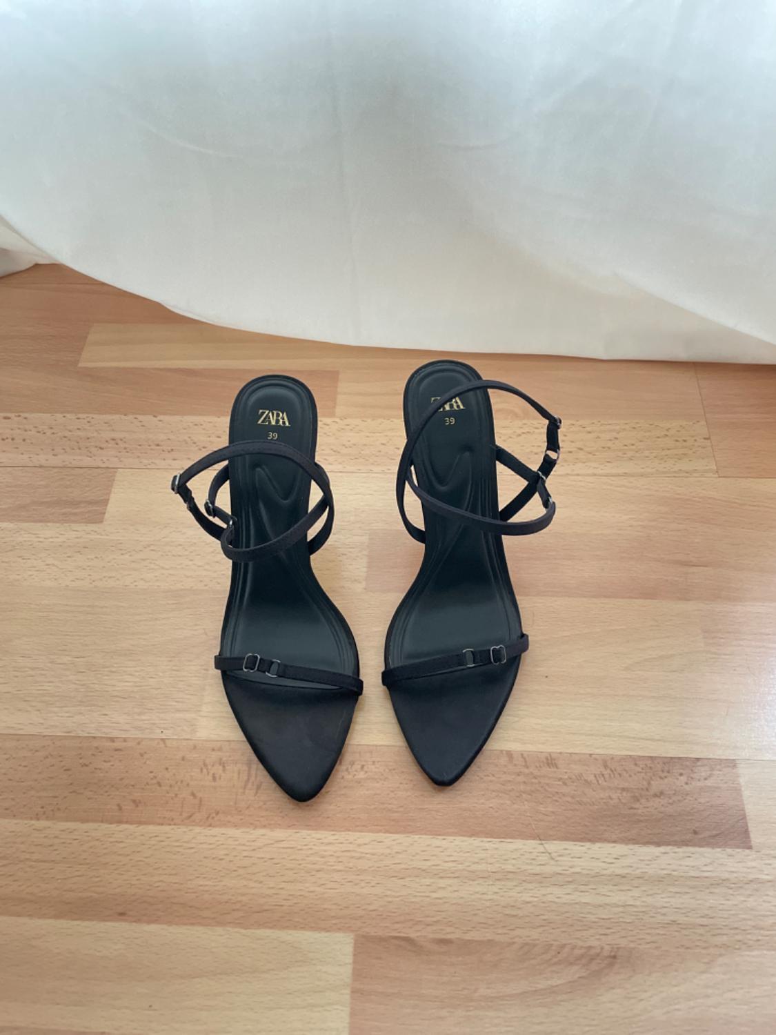 Black heeled sandals with thin crossed straps, on a light wooden floor. Visible text: "ZARA 39".