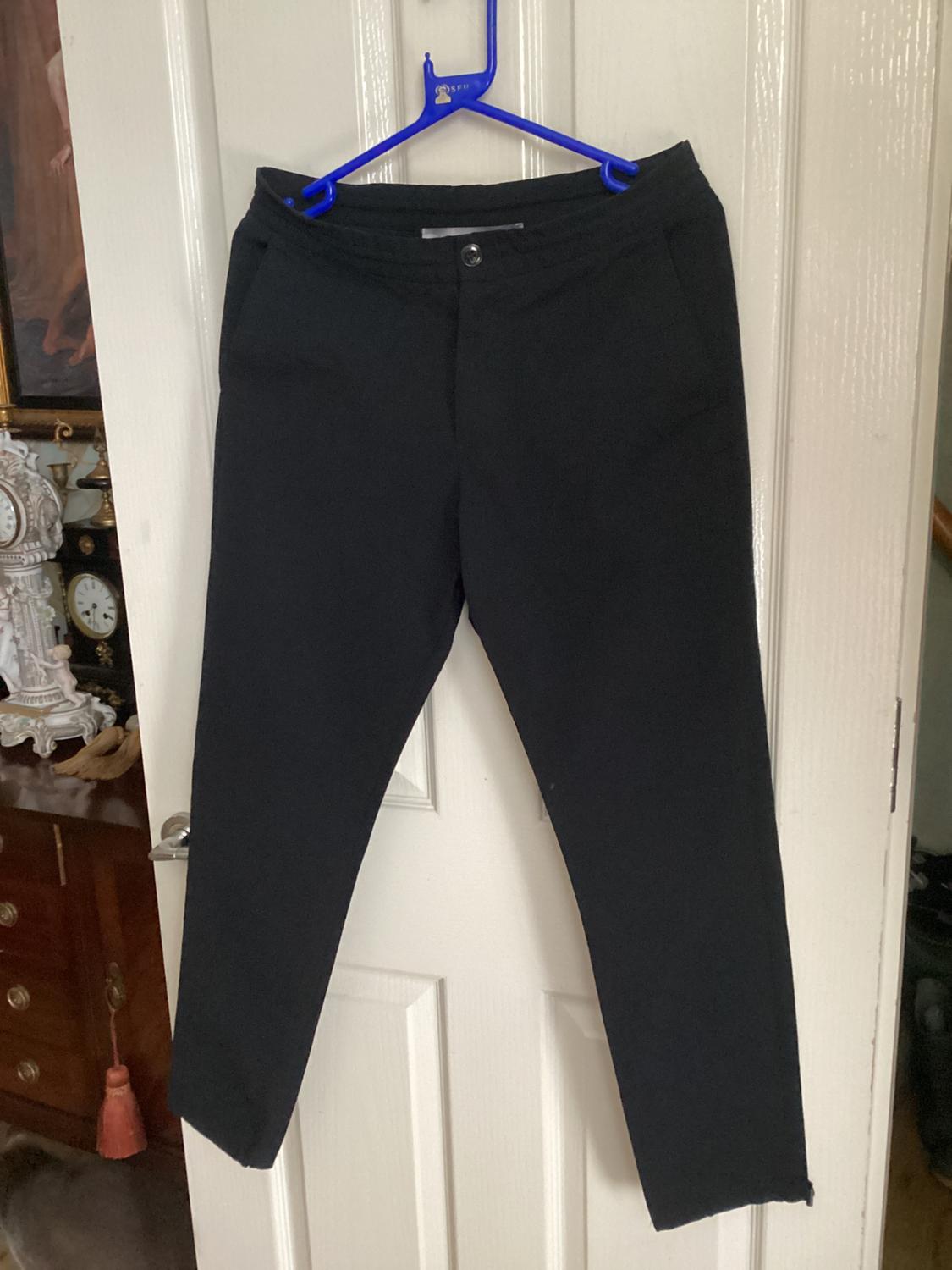 Front view of black trousers with side pockets and a front button hanging on a blue hanger.