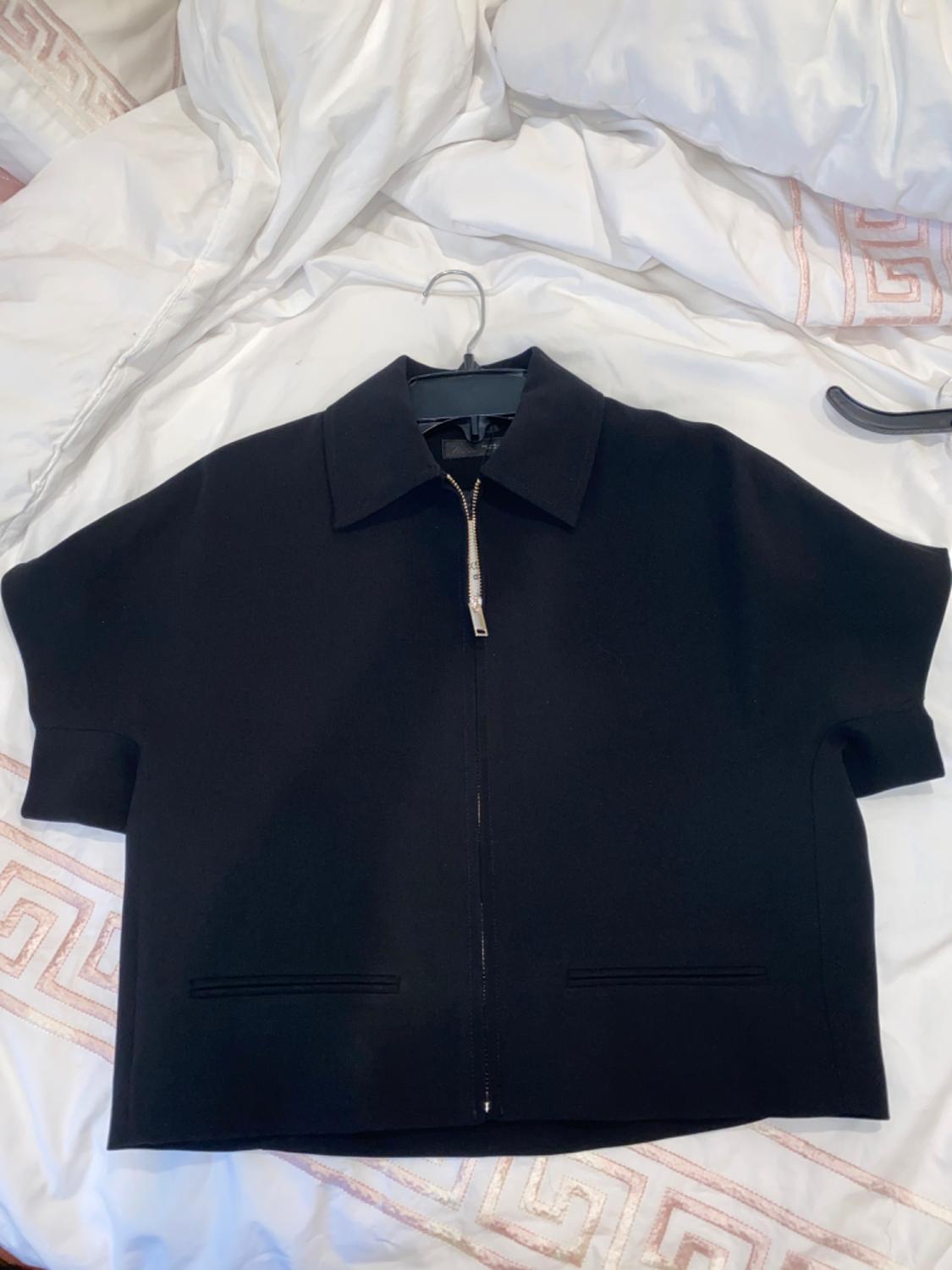 Black short-sleeved jacket with a front zip on a bed with white sheets and pink geometric details.