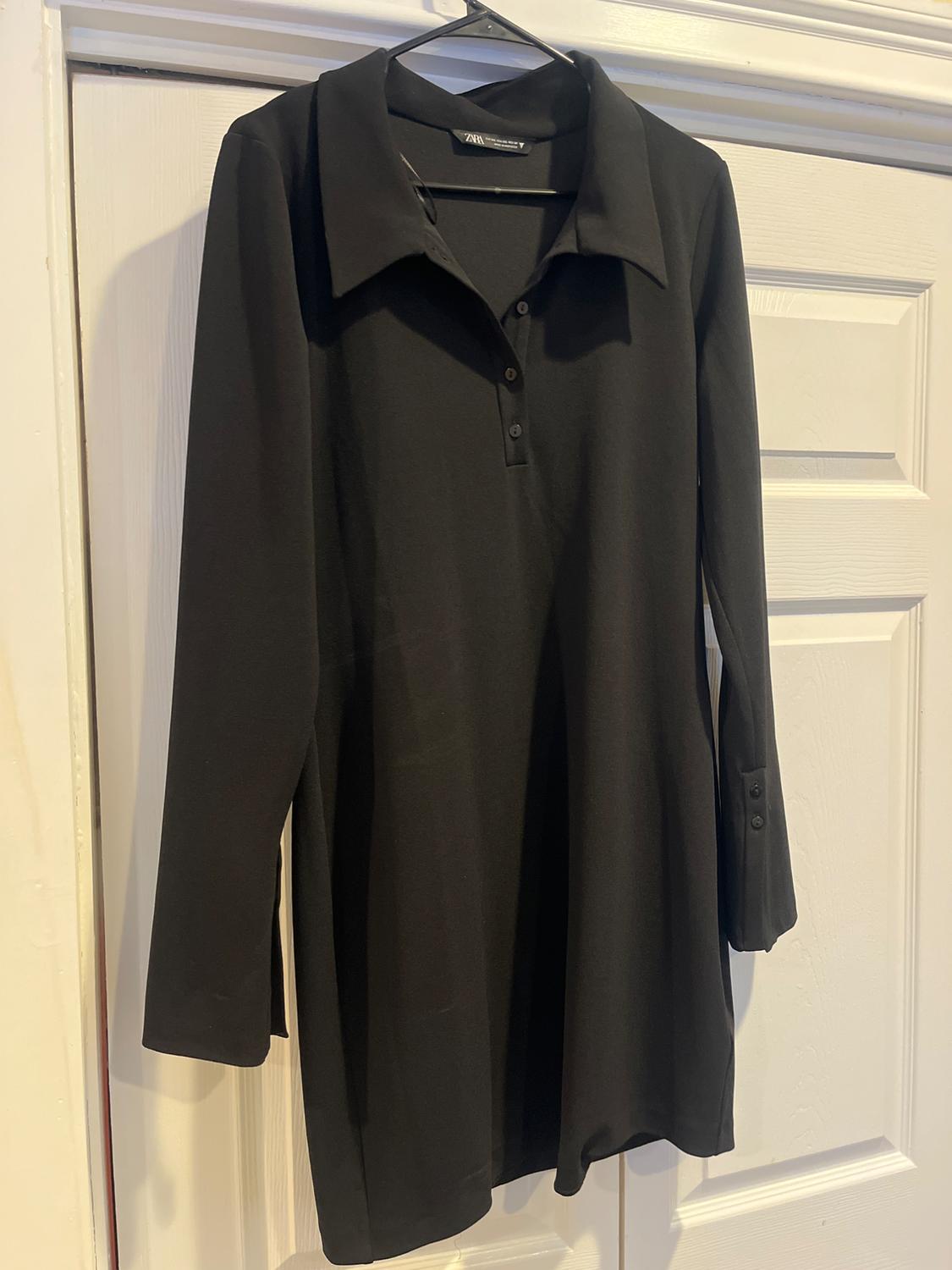 Black long-sleeved dress with a classic collar and buttons at the top, hanging on a hanger in front of a white door.