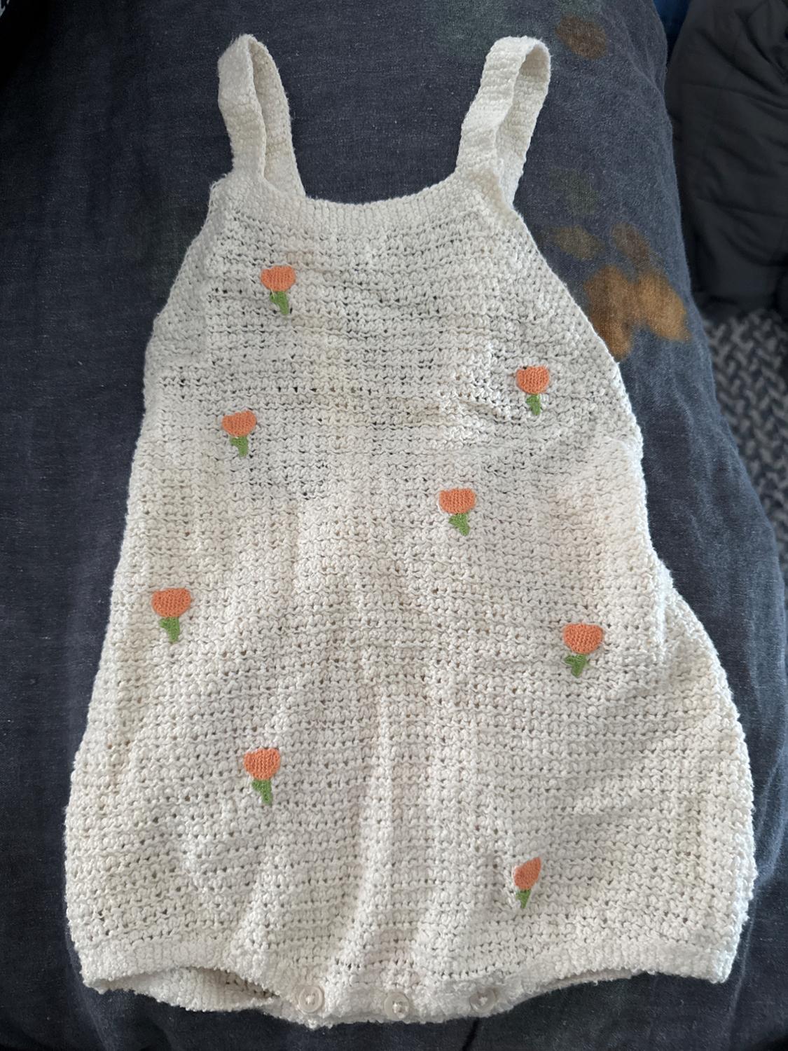 White woven jumpsuit with straps, decorated with small orange and green flowers. Dark background.