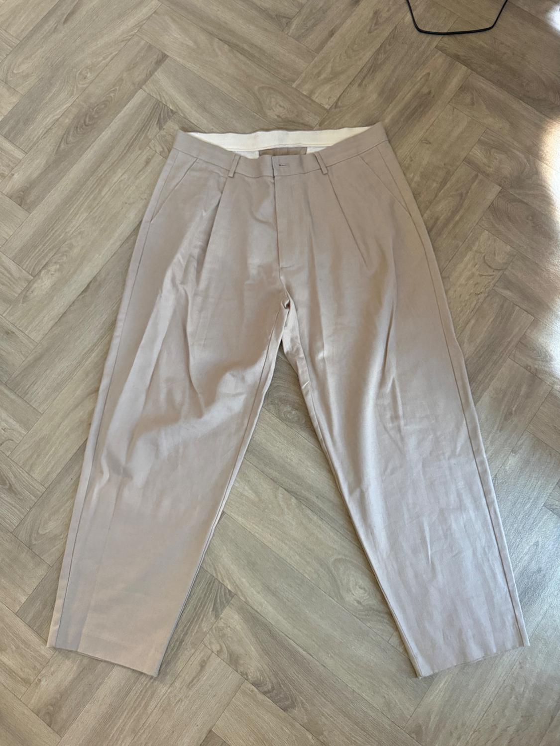 Beige pleated, loose-fit trousers, laid flat on a herringbone wooden floor. Casual style.