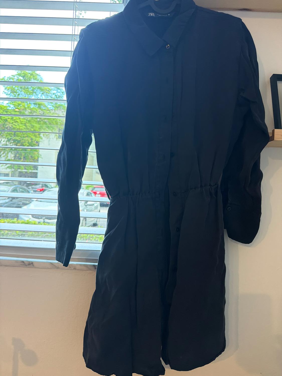 Black long-sleeved button-down shirt dress with ZARA label, hanging on a hanger next to a window with white blinds.