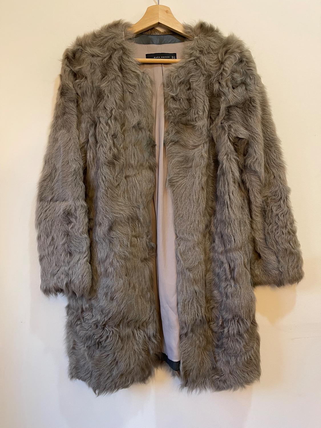Grey faux fur coat hanging on a wooden hanger. Visible label: "Zara Woman". White background.