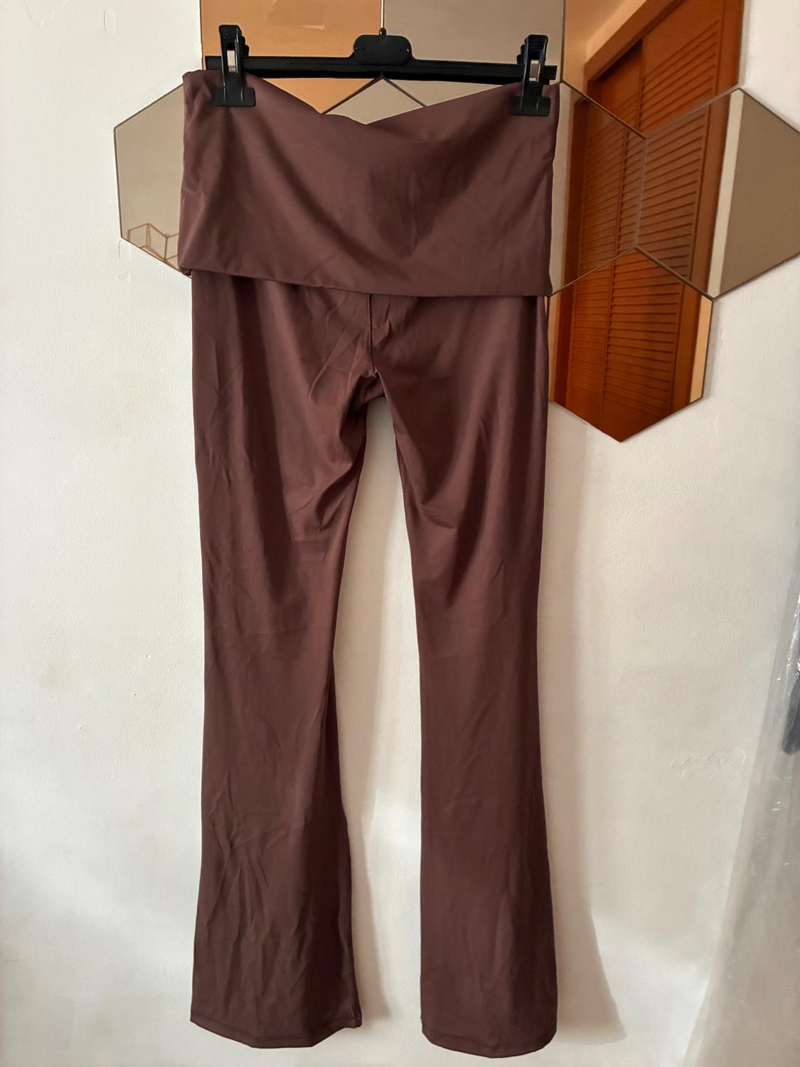 High-waisted brown trousers hanging on a black hanger, with a white wall background and hexagonal mirror.